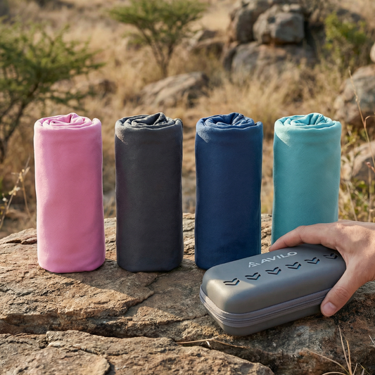 Four rolled-up towels in pink, gray, blue, and teal on a rock with a hand holding a device labeled 'AVILO' in a natural setting.