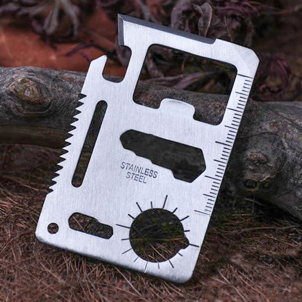 Stainless steel card multi-tool leaning against a weathered branch on a bed of pine needles