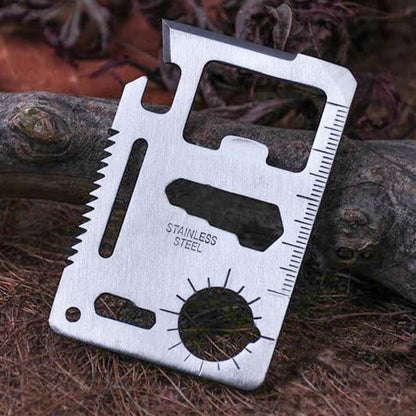 Stainless steel card multi-tool leaning against a weathered branch on a bed of pine needles