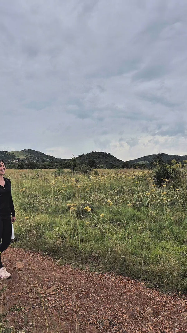 A woman walks a red dirt trail through highveld grassland in flower, hills behind her, flask in hand.