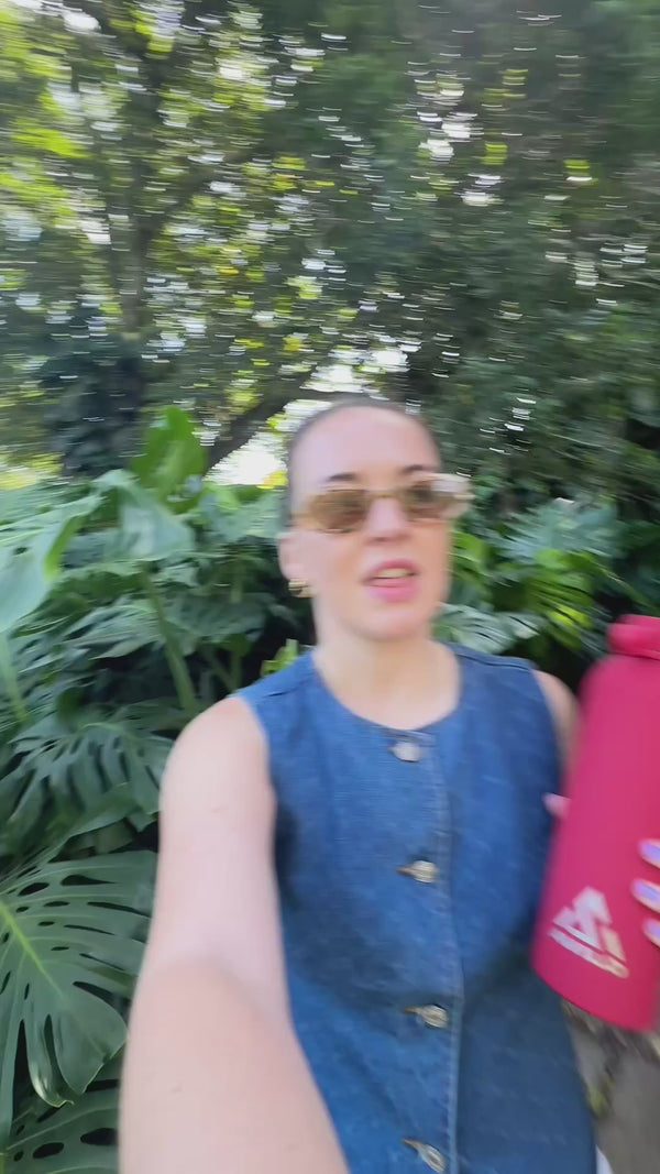 A woman walks through a lush garden carrying a red Avilo flask.